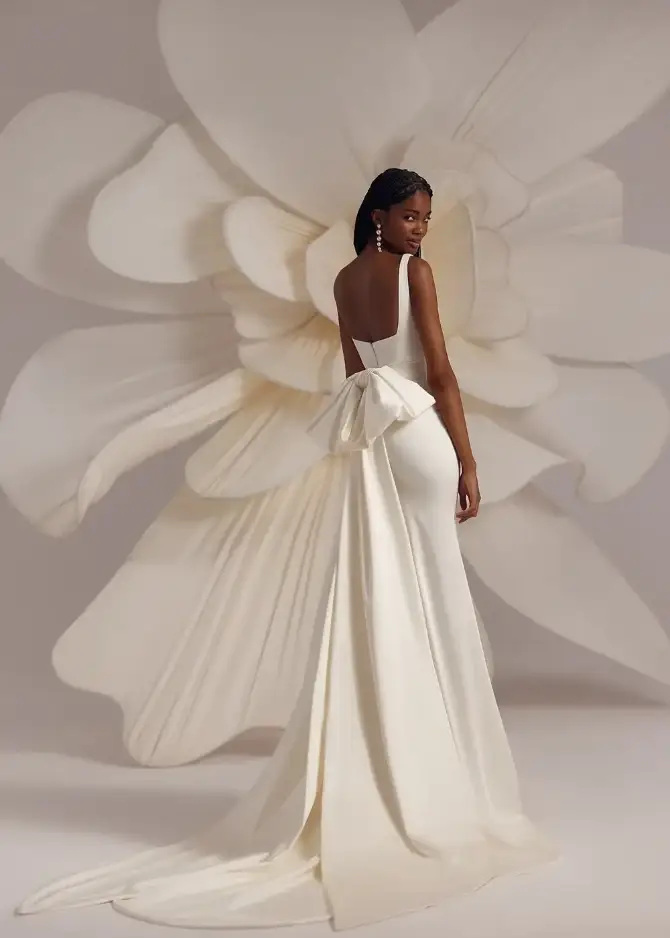 Model is wearing a fitted gown with a square neckline, long skirt, detachable oversized bow at the back, and a floral-edged veil.