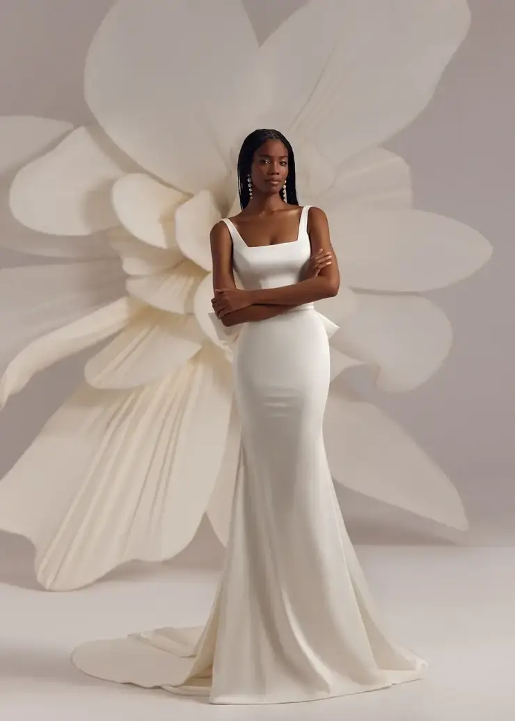 Model is wearing a fitted gown with a square neckline, long skirt, detachable oversized bow at the back, and a floral-edged veil.