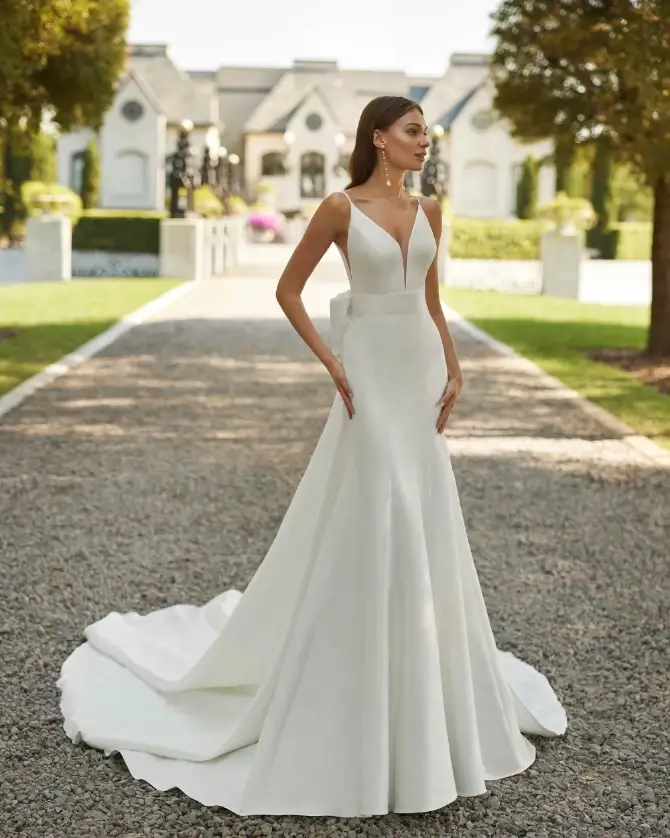 Bride in a white, elegant wedding dress stands outdoors on a gravel path, with a mansion and manicured garden in the background, exuding grace.