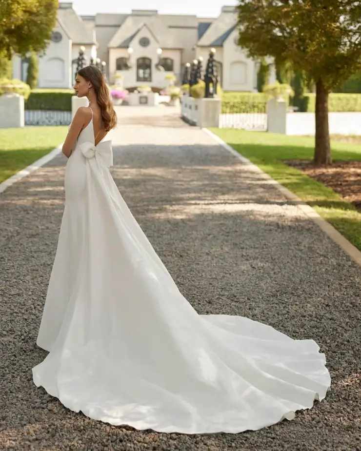 Bride in elegant white gown with long train and bow detail, standing on a gravel path leading to a grand estate. Serene, romantic atmosphere.