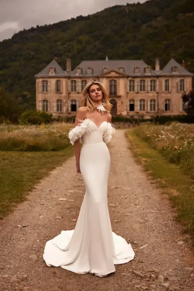 A woman in an elegant, off-shoulder white gown with feather details stands on a dirt path. She is in front of a grand, vintage mansion surrounded by lush greenery. The mood is romantic and serene.