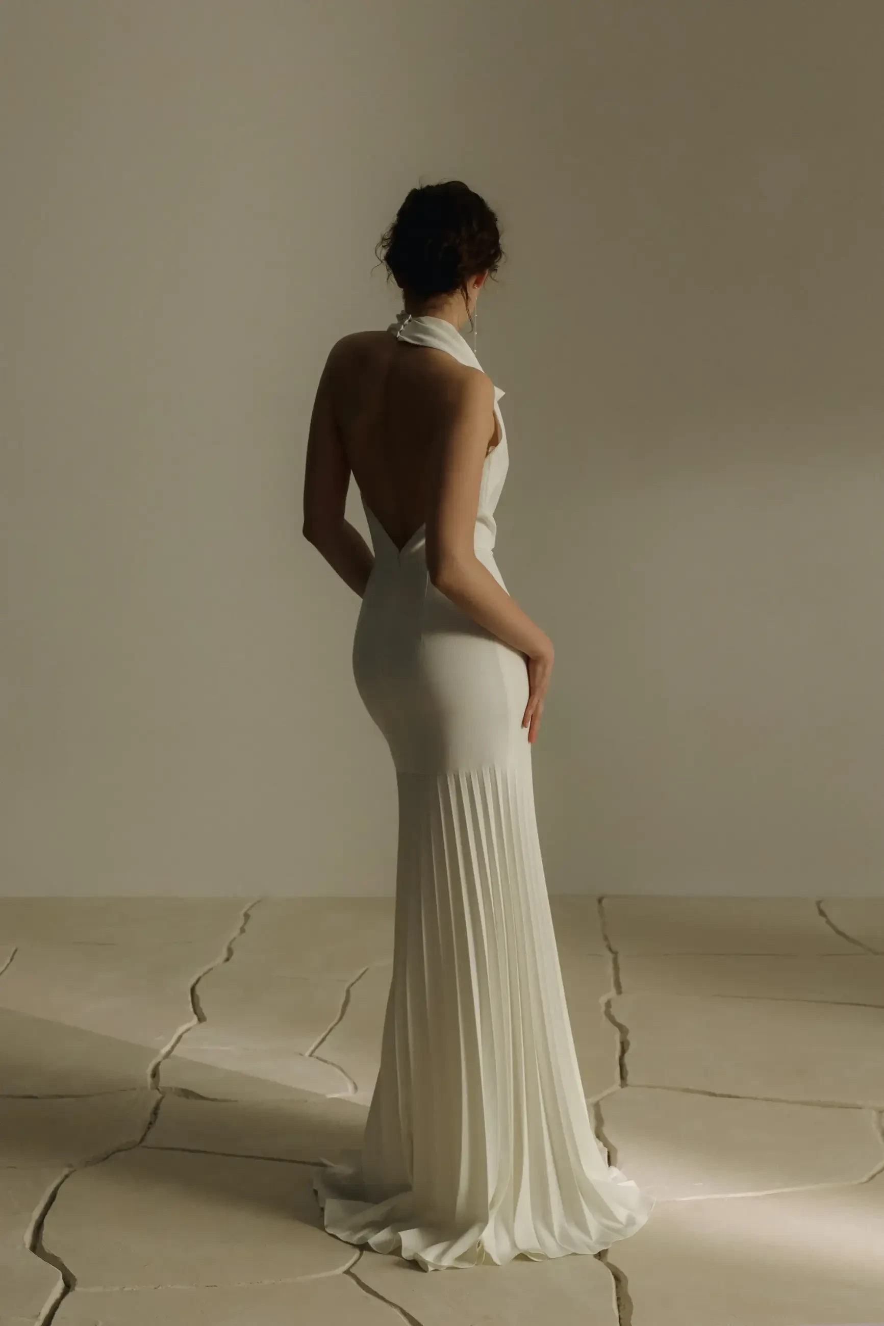 A woman wearing a backless, white gown standing with her back to the camera, on a textured surface.