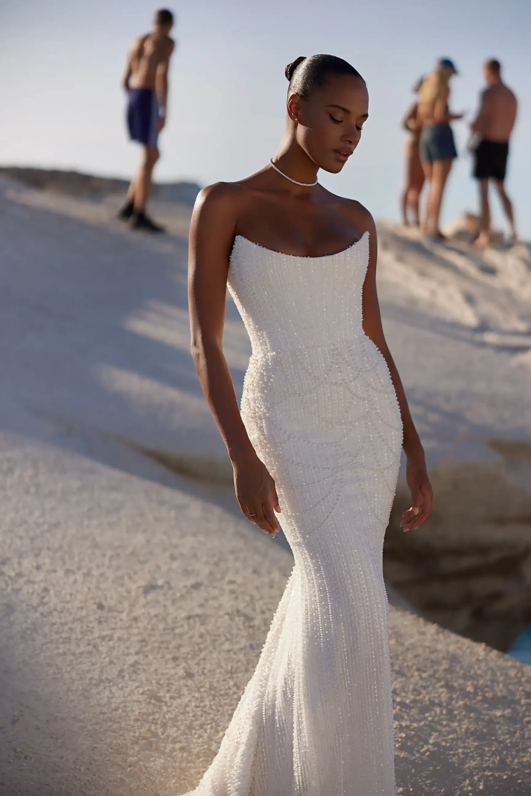 A model in a fitted, elegant white gown stands gracefully on a sandy surface with ocean waves in the background.