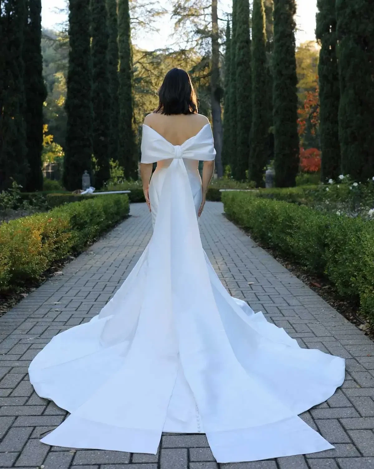 Bride in an elegant white gown with a long train and bow walks along a garden path lined with tall trees, creating a serene and picturesque scene.