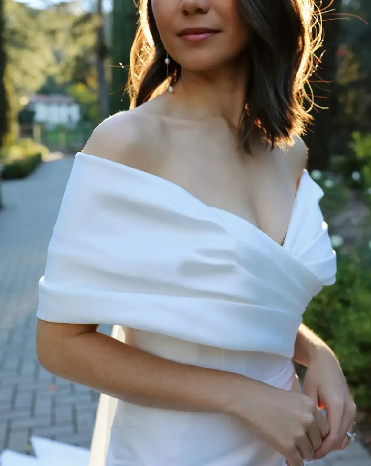 A woman in an elegant off-shoulder white dress stands outdoors in soft sunlight, surrounded by greenery. The setting conveys a serene and romantic mood.