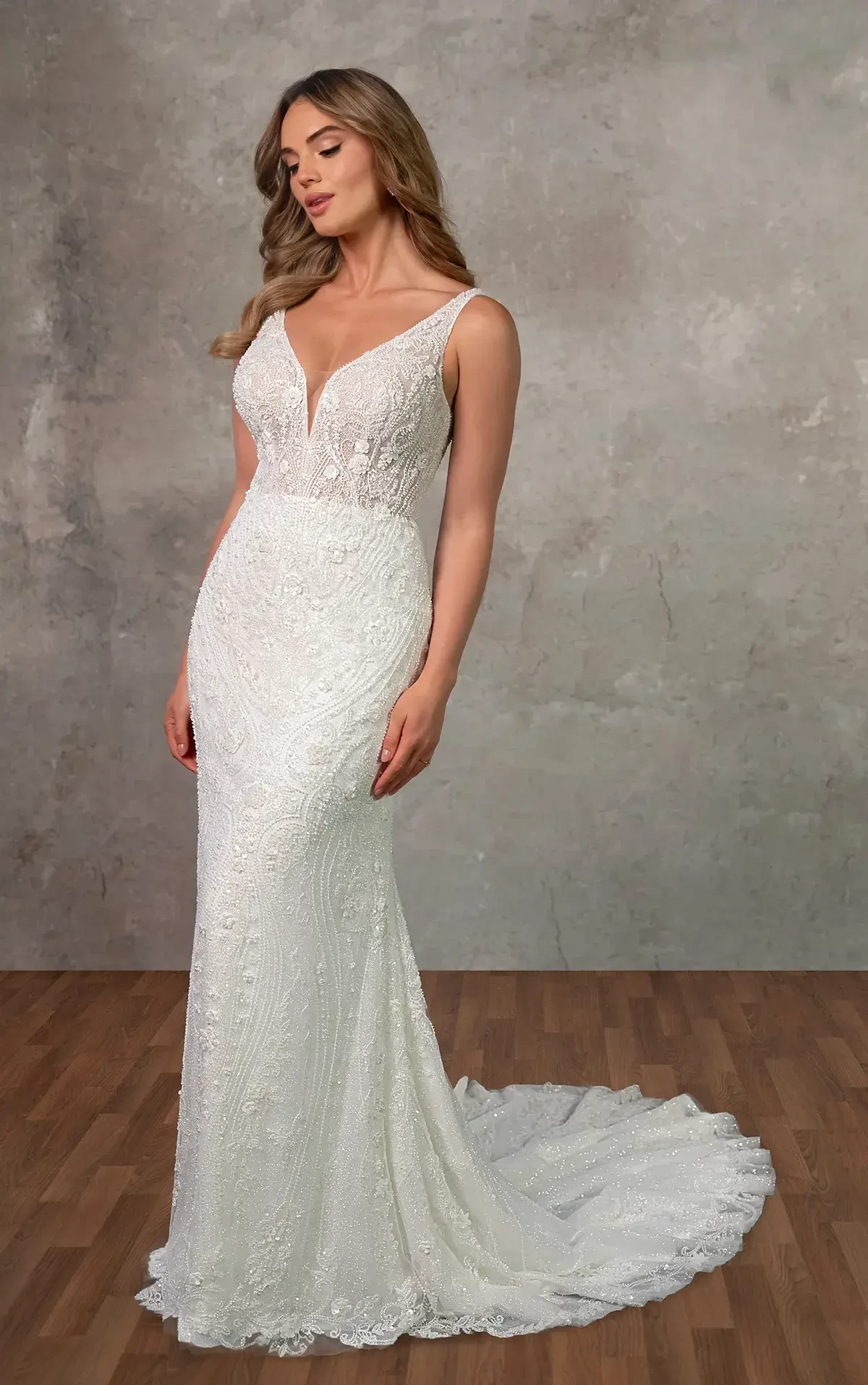 A woman wearing a fitted white wedding dress with intricate designs and a long train, posing gracefully against a simple background.