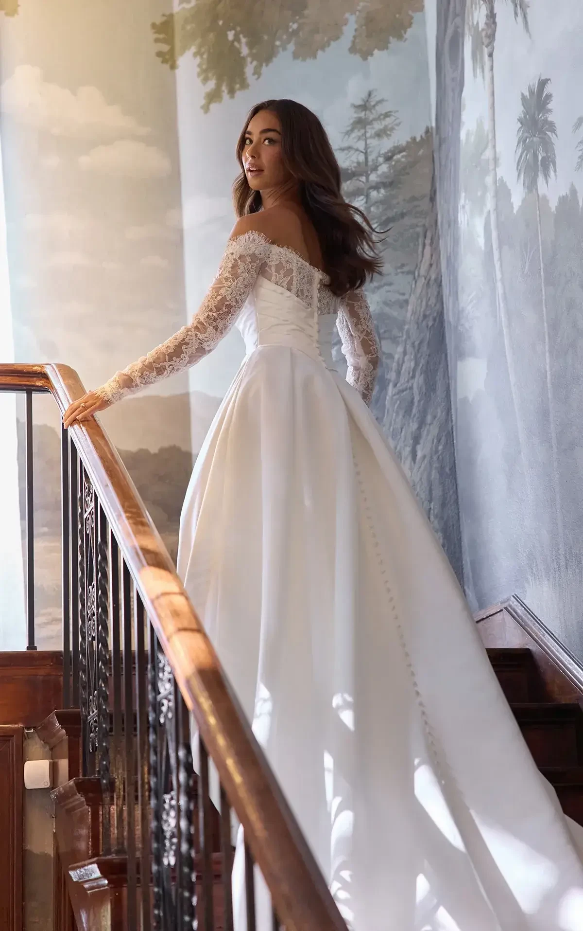 Bride in an elegant white gown with lace sleeves ascends a wooden staircase, surrounded by mural walls depicting trees, exuding grace and elegance.