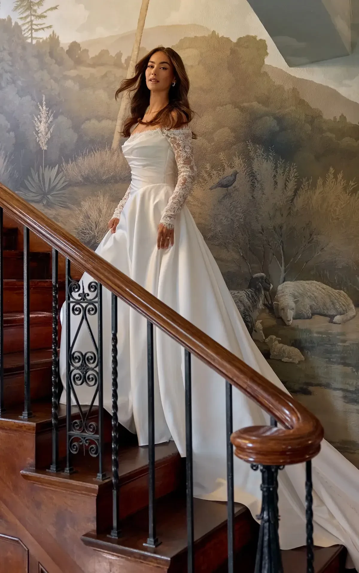A woman in a white wedding dress is standing on a staircase, with a painted mural of nature in the background.