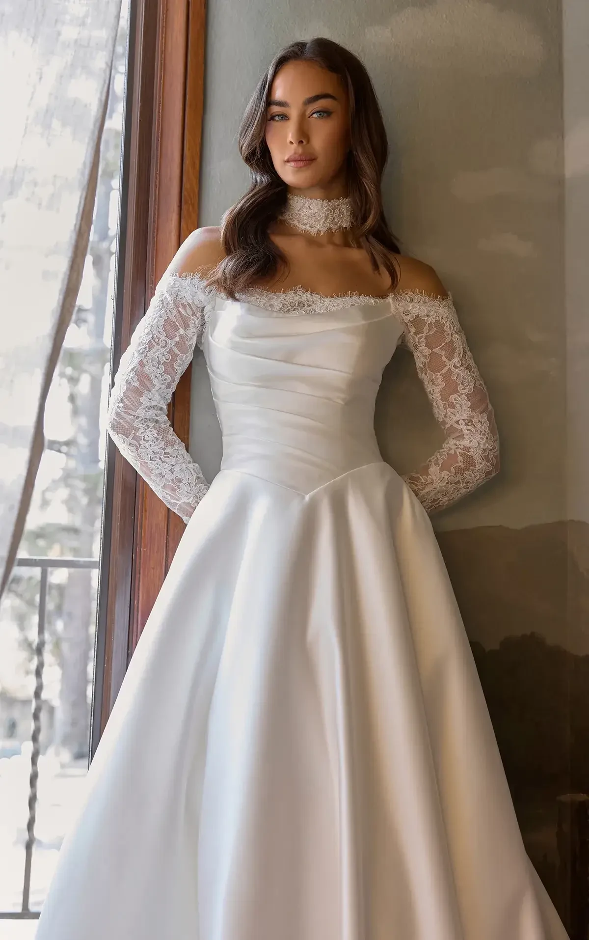 Elegant woman in a satin wedding dress stands by a window. The dress features lace sleeves and an off-the-shoulder design. She exudes calm confidence.