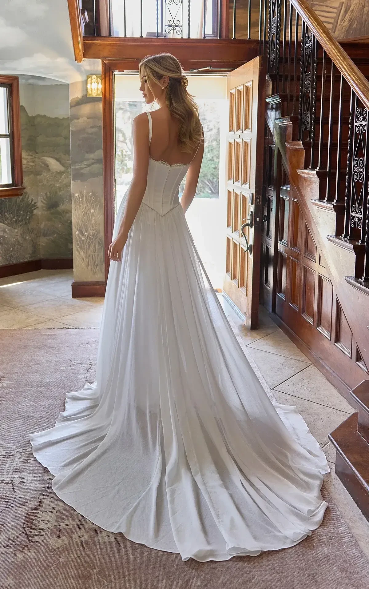 A woman in a flowing white wedding dress stands in a warmly lit, rustic hallway. Natural light streams through the door, creating a serene and elegant atmosphere.