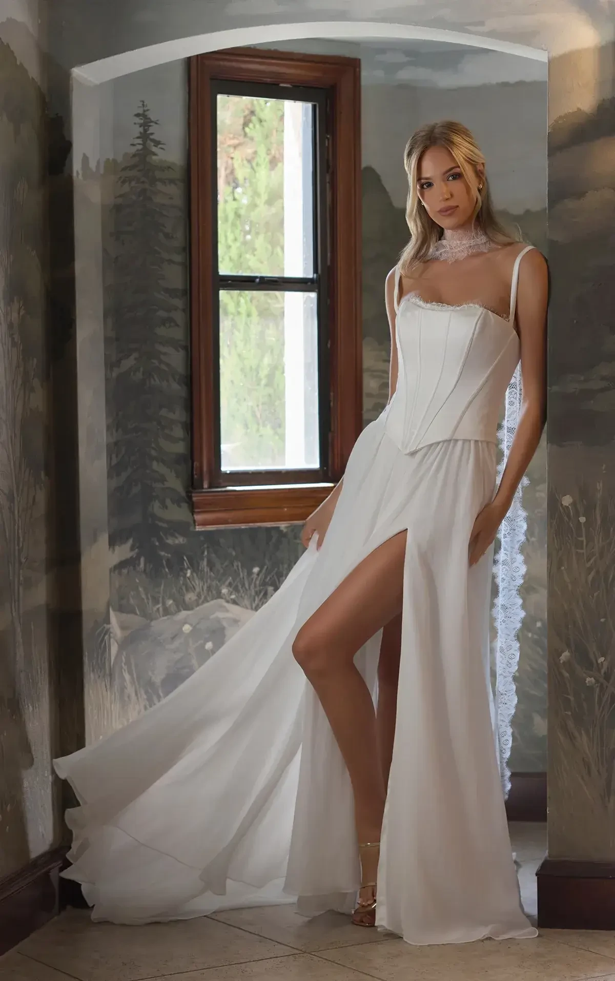 Woman in elegant white dress with a high slit stands in a mural-painted corridor. Her expression is serene, framed by a small window and soft natural light.