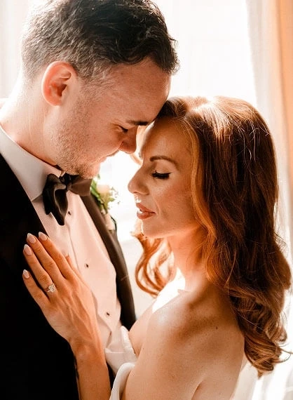 Сouple wearing a white gown and a black suit near the window