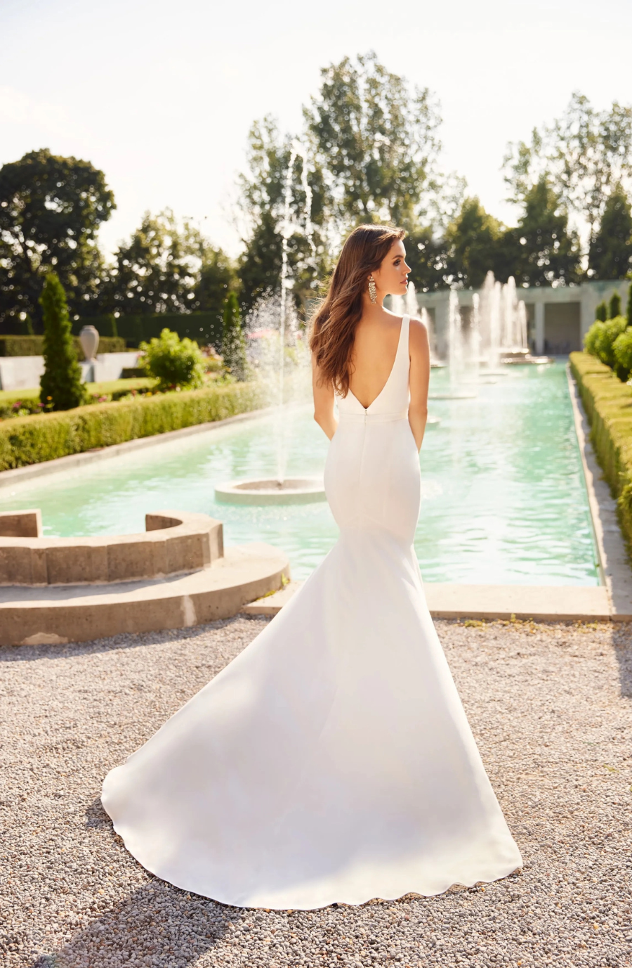 Bridal scene with woman in a fitted, backless white wedding gown, standing near a serene fountain. The atmosphere is elegant and tranquil, set in a lush garden.